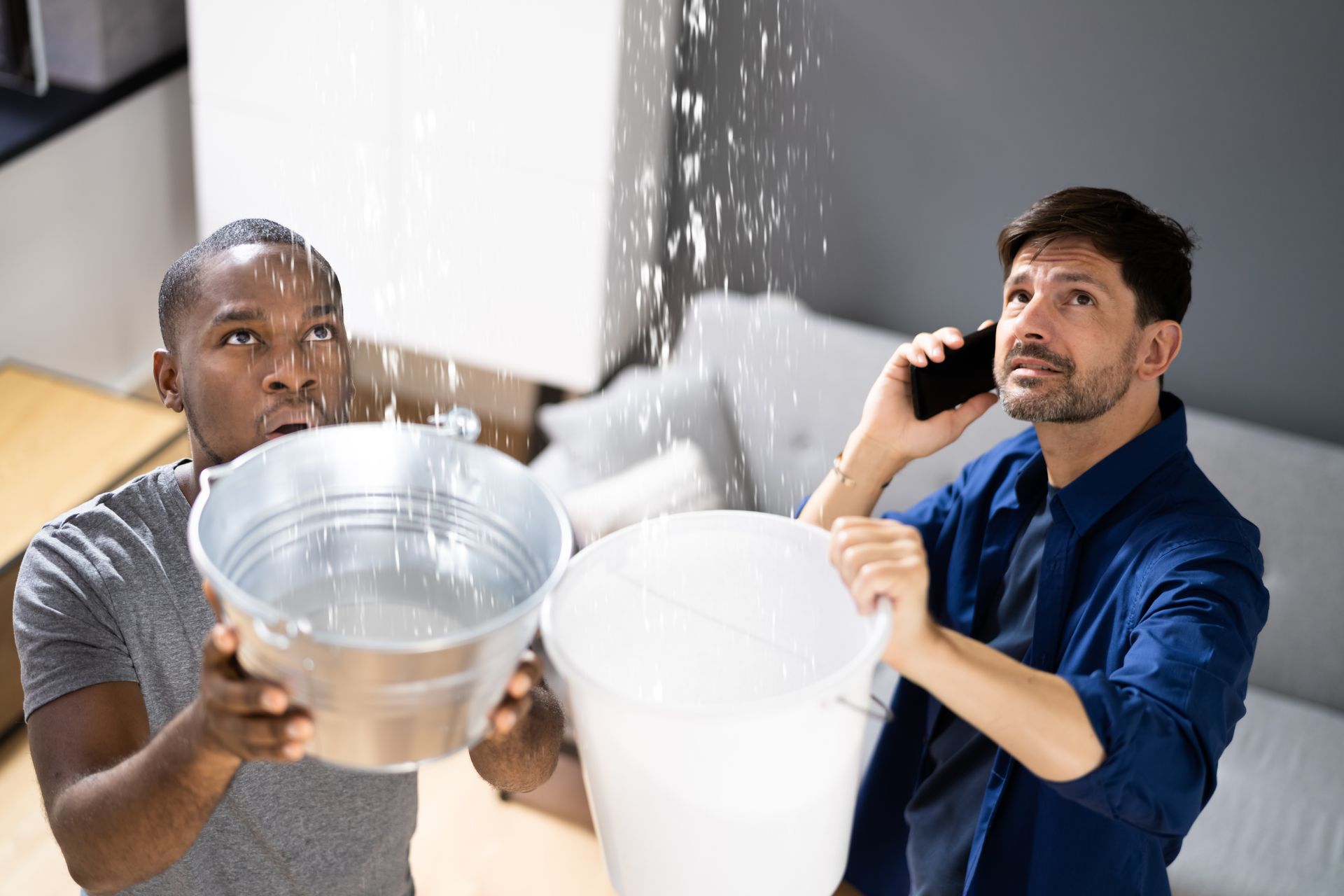 Two men catch water leaking from a ceiling into buckets, one holding a phone in a flooded room