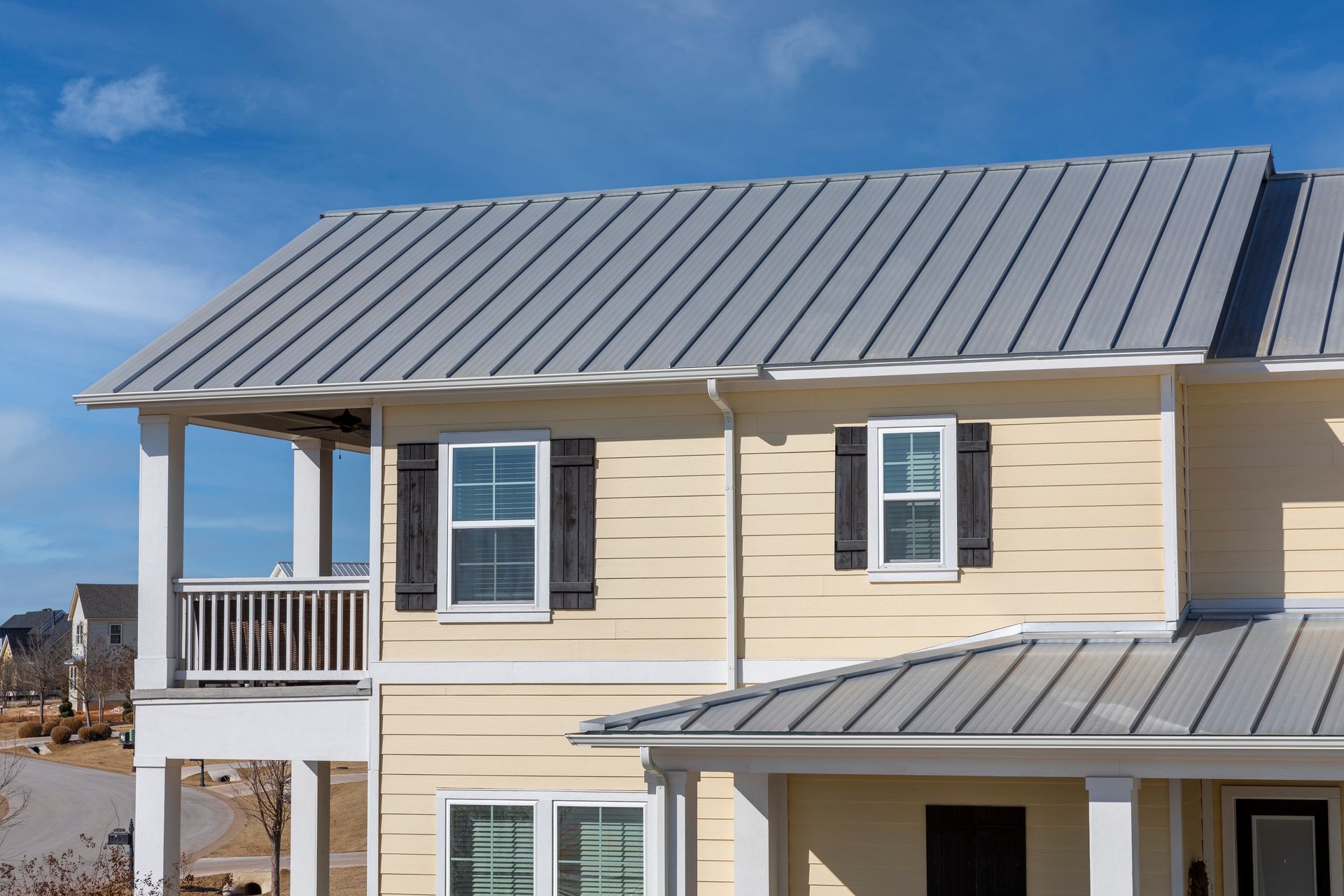 Two-story beige house with a gray metal roof and white trim under a blue sky
