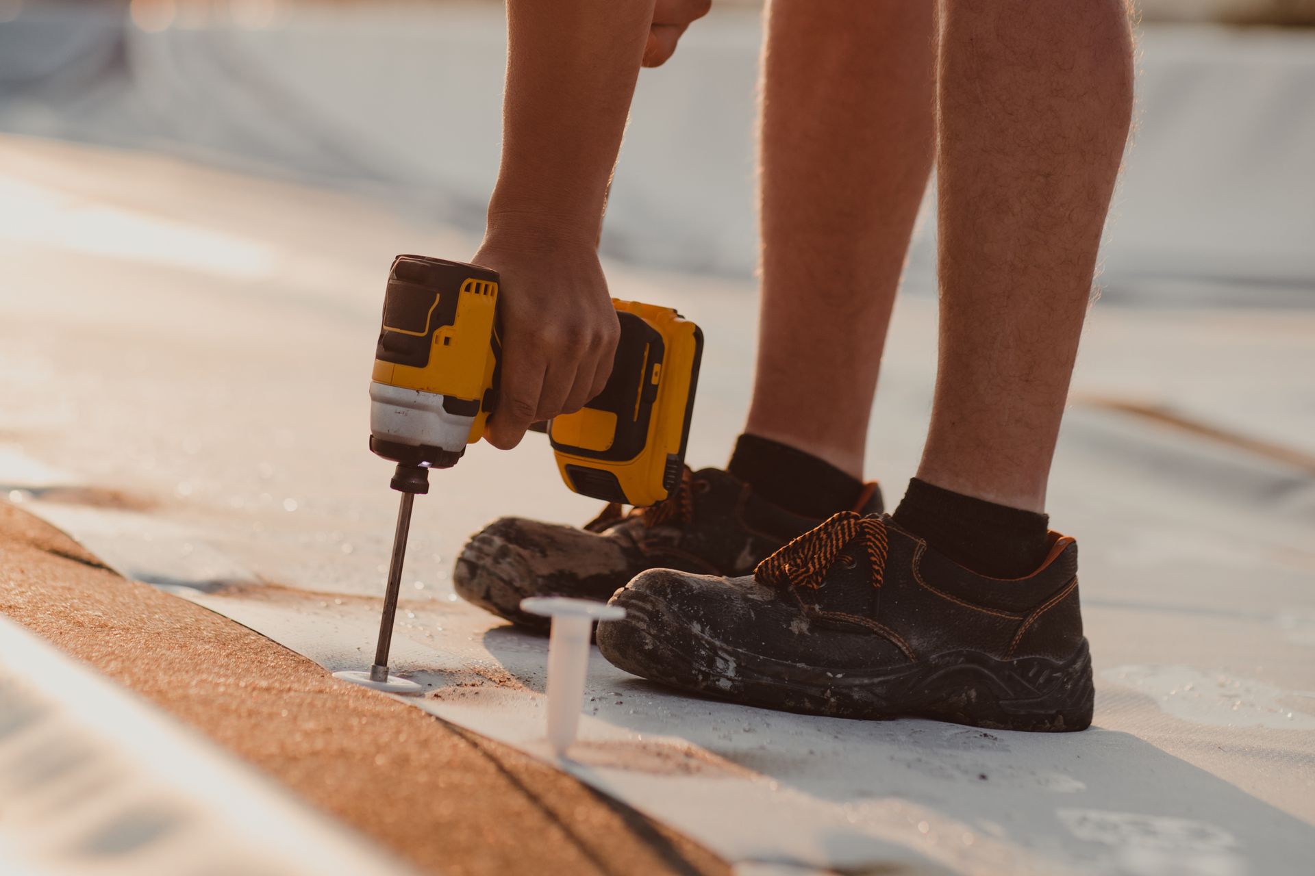 Worker drilling a roof membrane with a yellow power drill, wearing black work shoes and shorts
