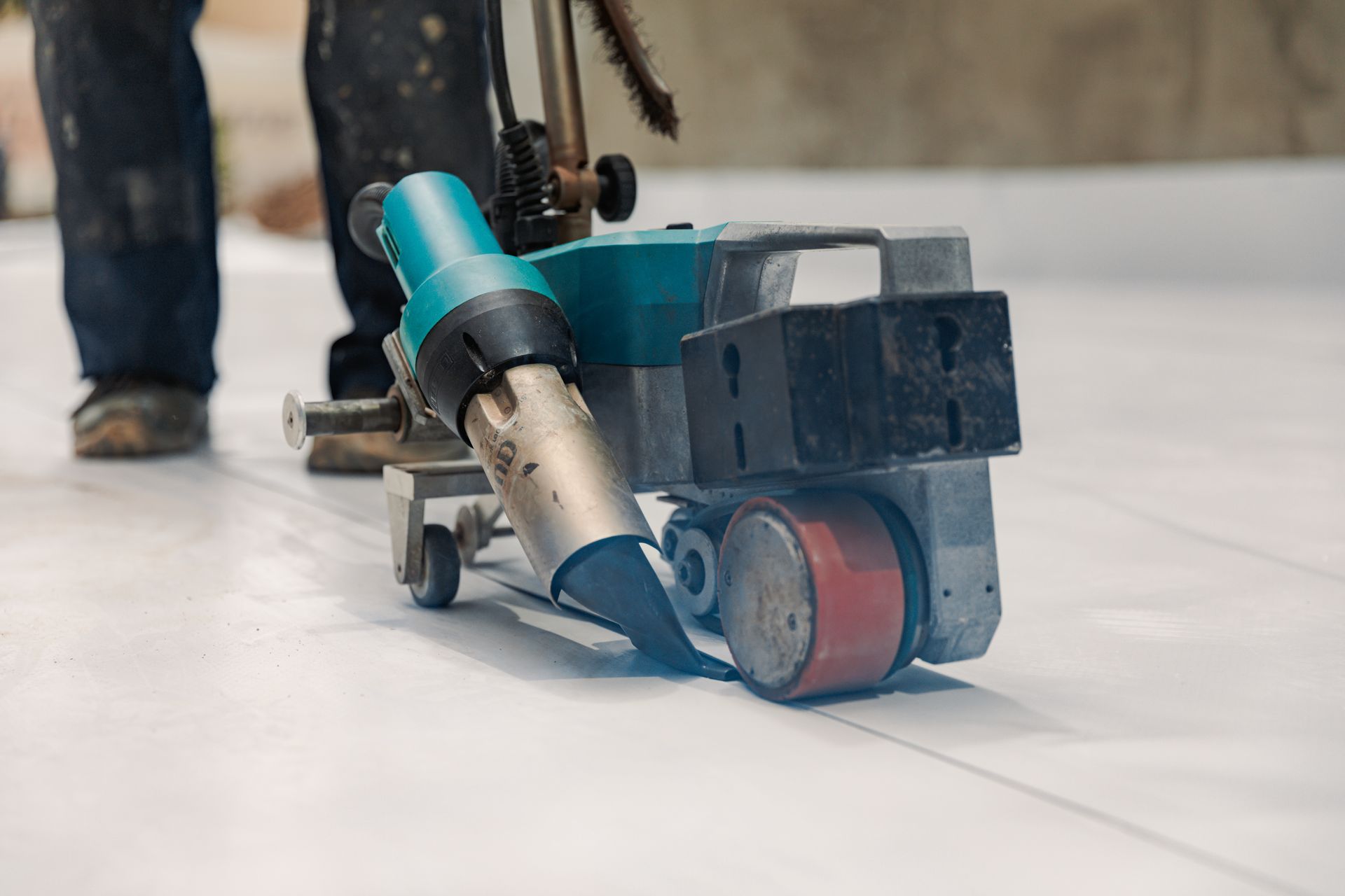 Worker using a floor grinder with a blue hose on a tiled floor, creating dust near a wall.
