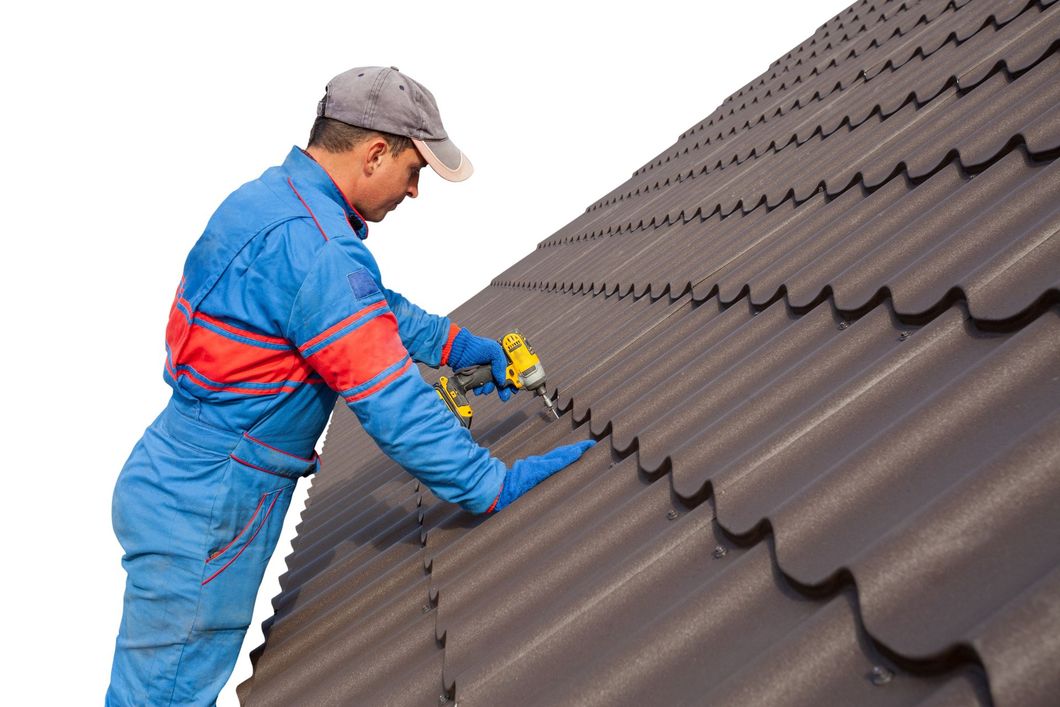 Worker in blue coveralls installing brown roof tiles on a steep roof using a power drill