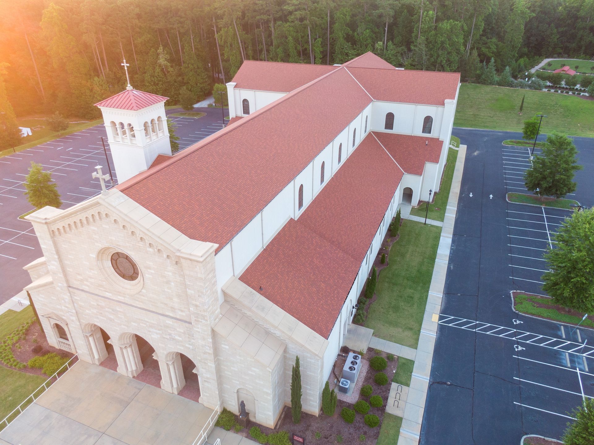 Aerial view of a white stucco church with a red tile roof beside an empty parking lot and trees.