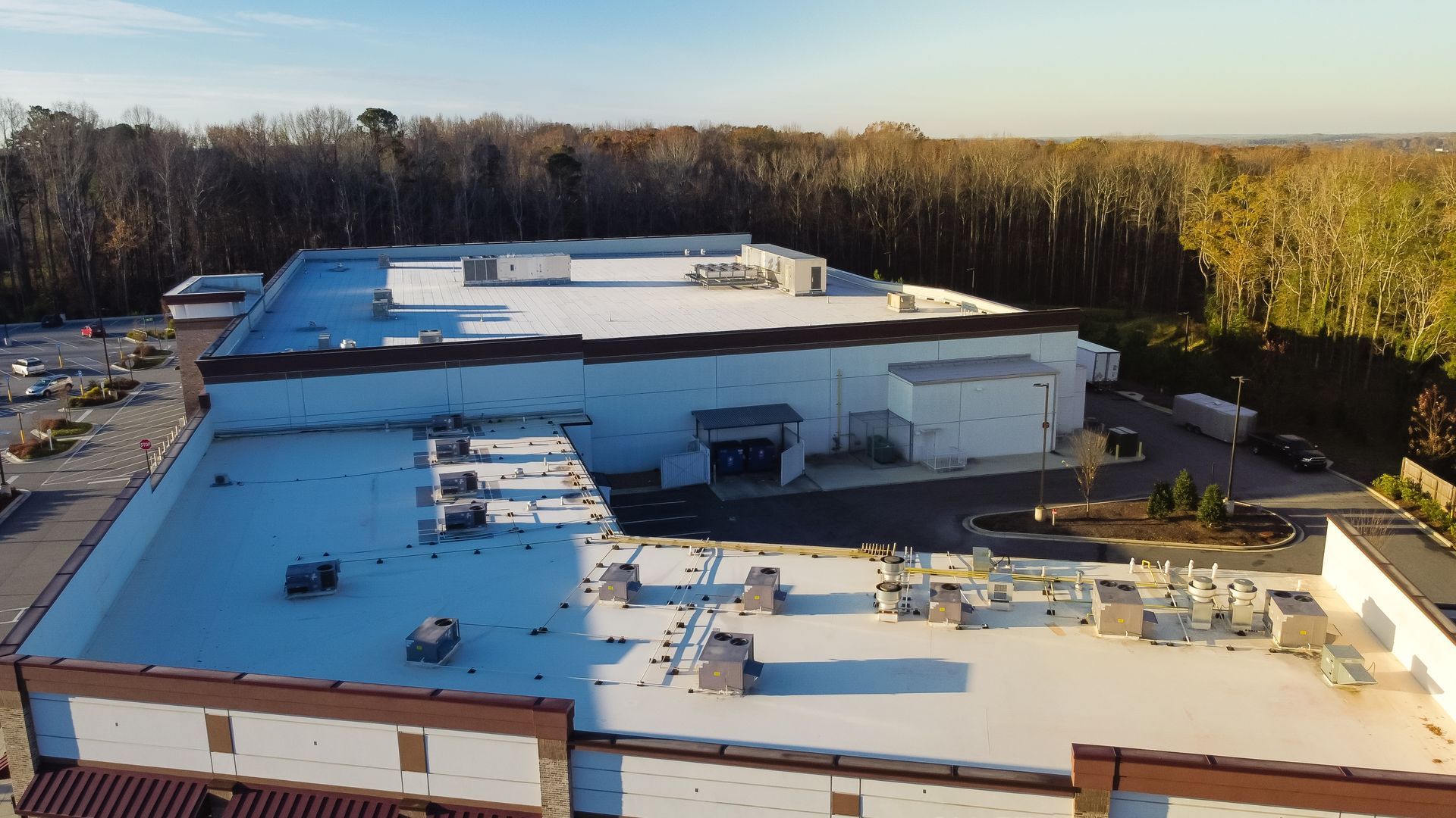 Aerial view of a large white industrial building with a flat roof beside a wooded area at sunset