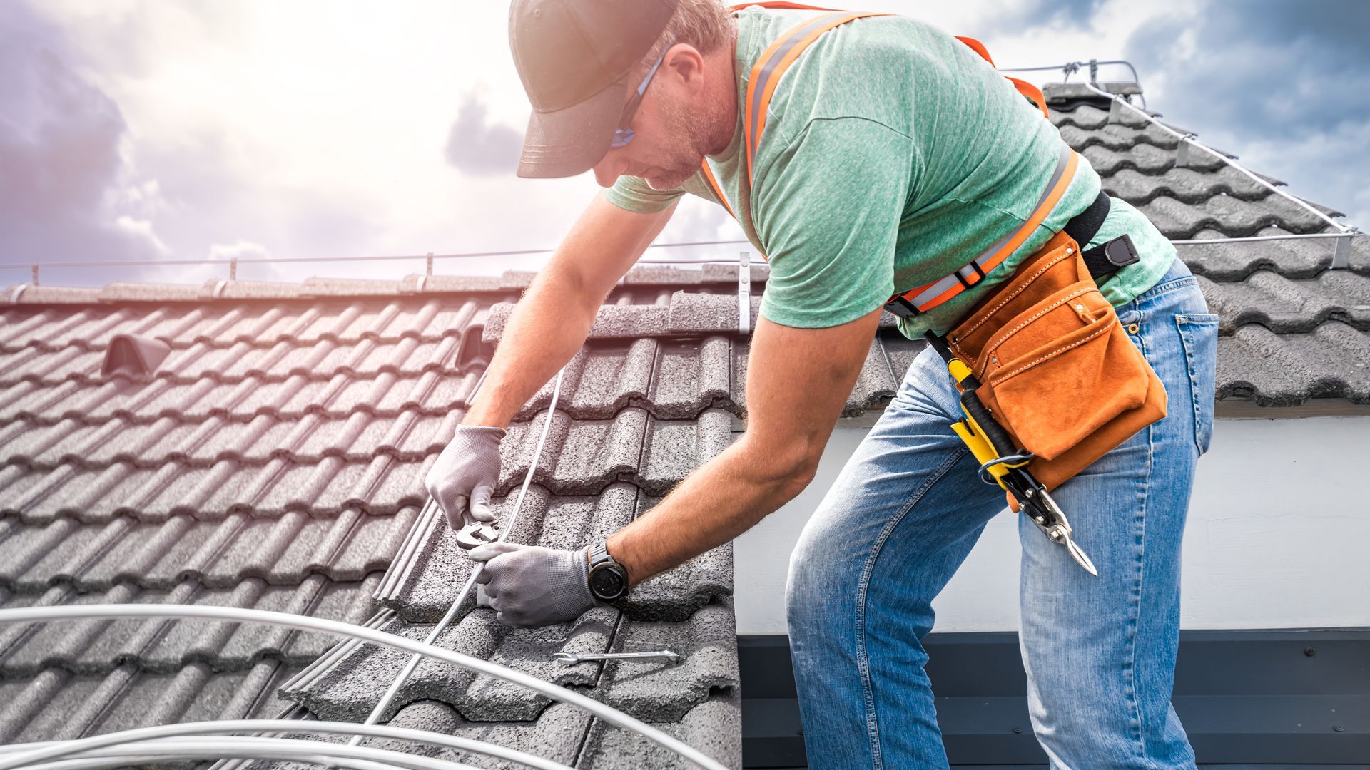 Worker installing roof tiles with safety gear and tool belt on a gray tiled roof.