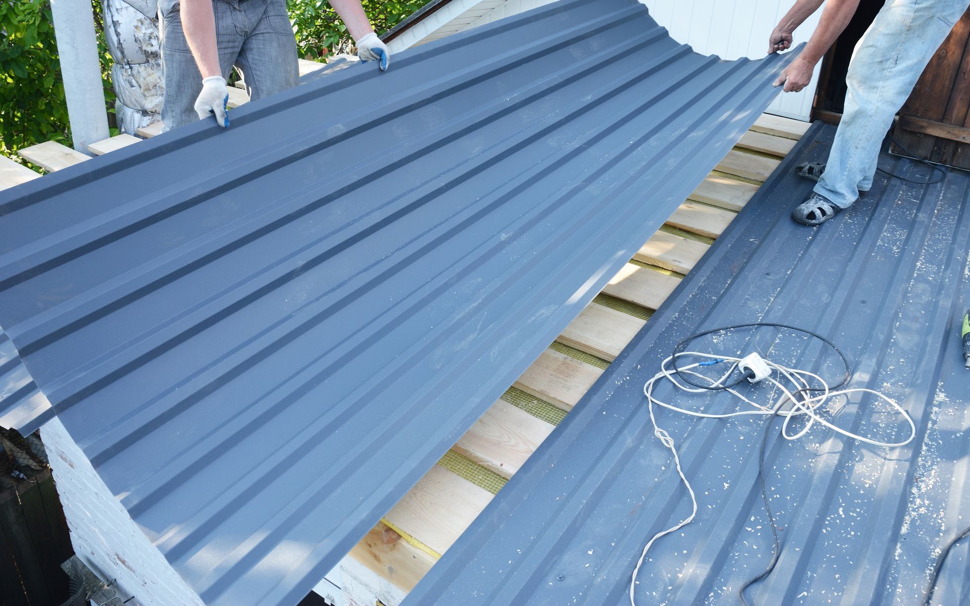 Workers installing blue corrugated metal roofing on a roof frame, with tools and cables nearby