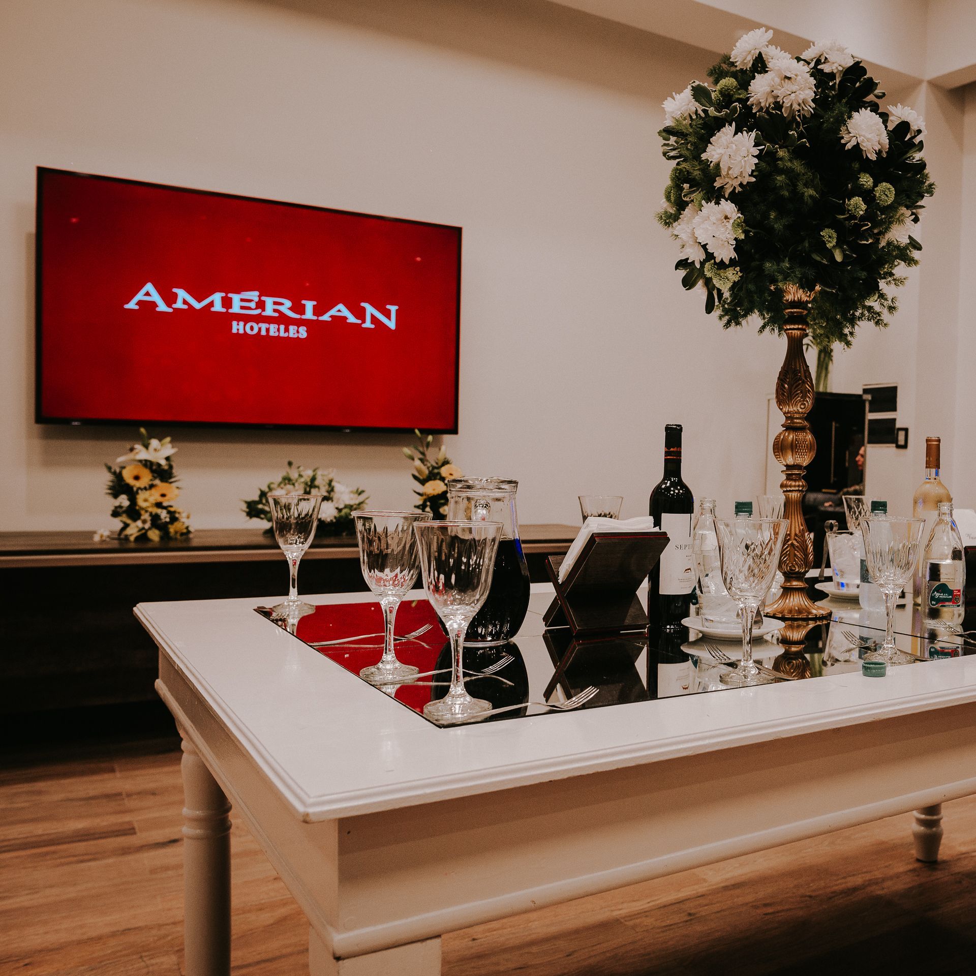 A table with wine glasses and bottles in front of a television that says amerian hotel