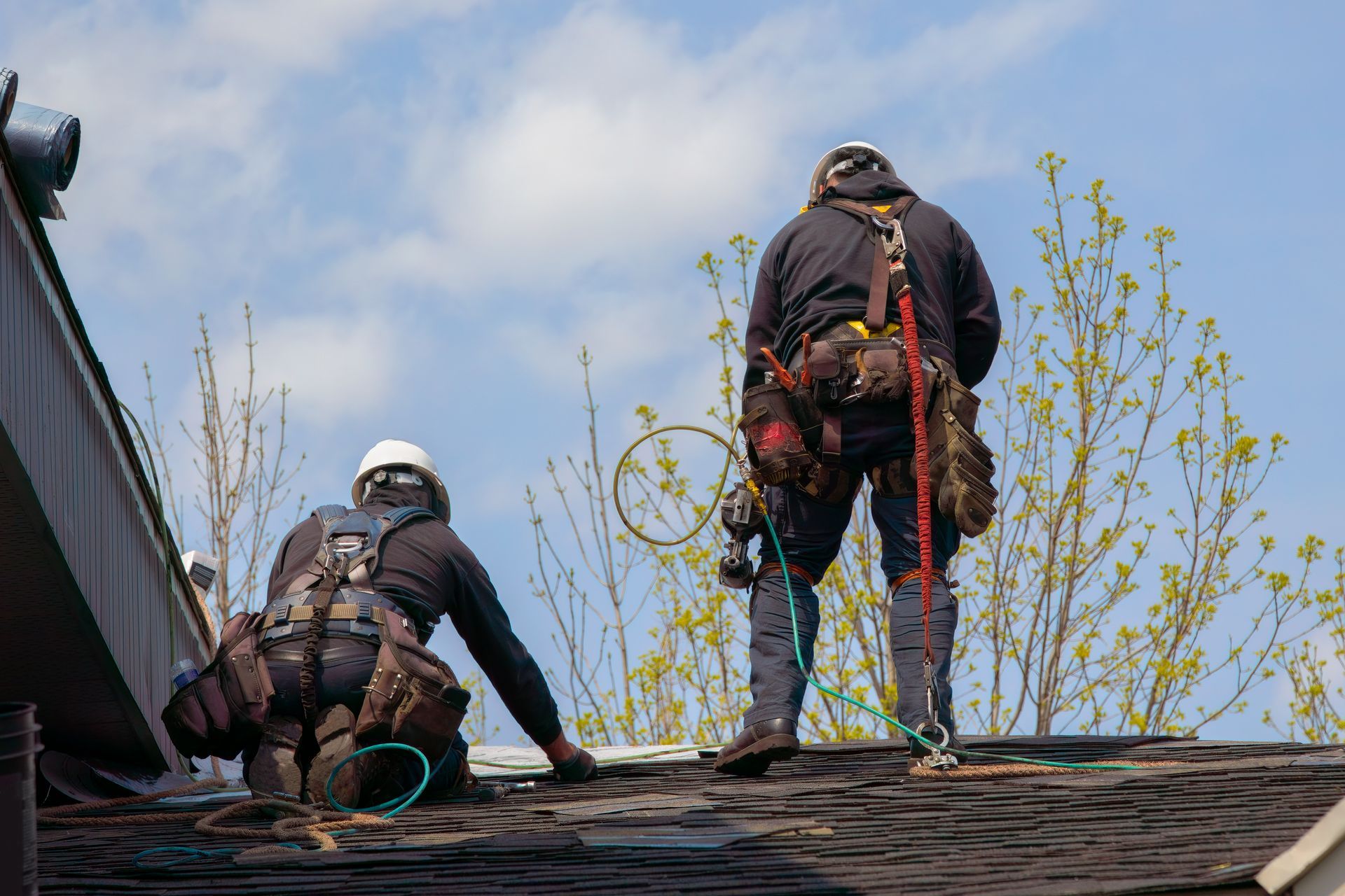 Two roofers wearing safety harnesses on a rooftop, one kneeling, the other standing, with a partly cloudy sky in the background.