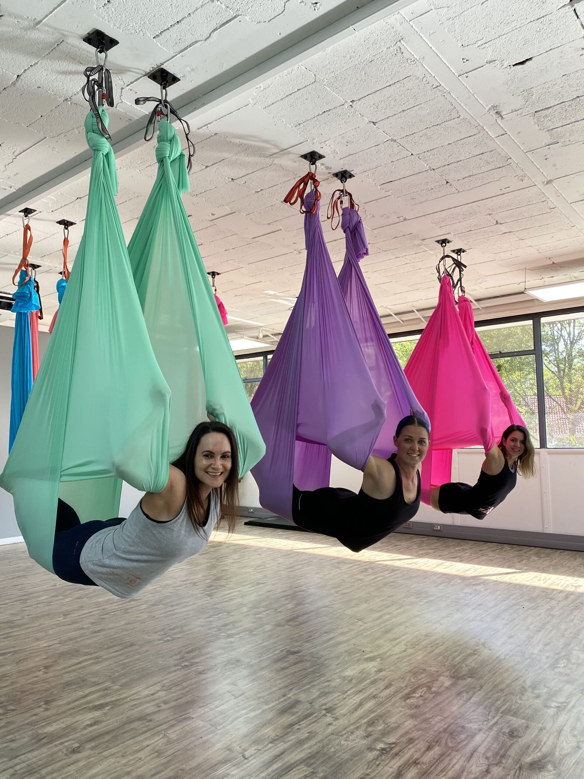 Three individuals suspended in colorful aerial silks, performing poses while lying on their backs in a spacious indoor setting.
