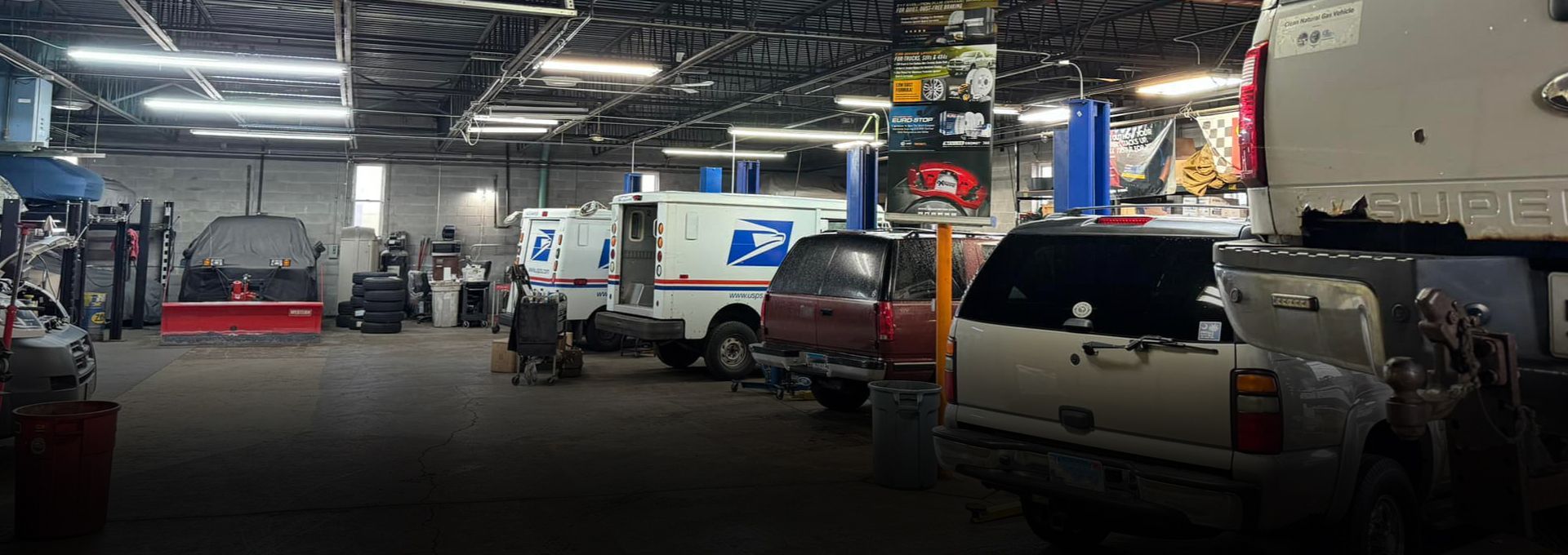 Inside a busy auto repair shop with multiple vehicles, a mix of equipment, and bright overhead lights | S&C Automotive Inc