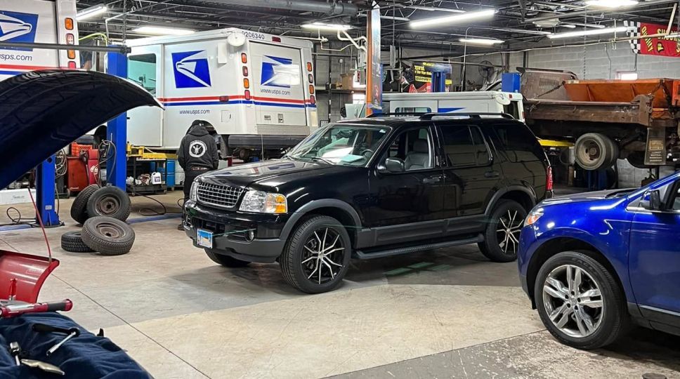 Black SUV in a repair shop with a USPS truck in the background, another car on the right, and tools visible | S&C Automotive Inc