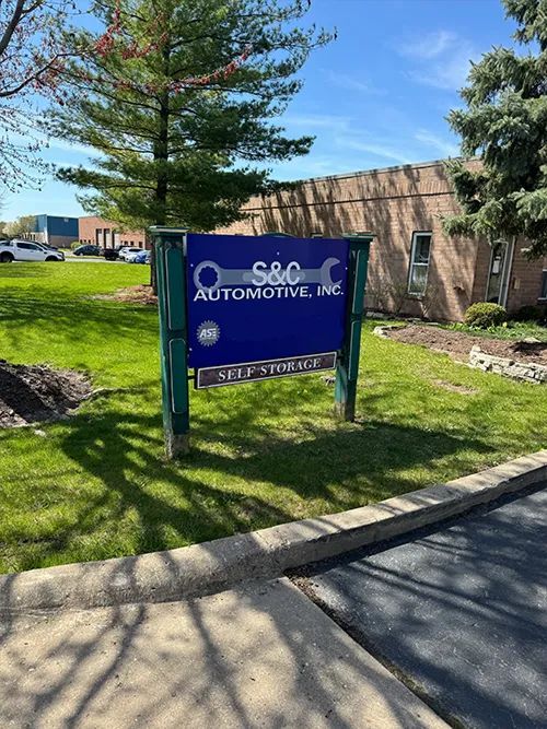 Sign for S&C Automotive, Inc., blue and white on green, with phone number, in front of building and lawn | S&C Automotive Inc
