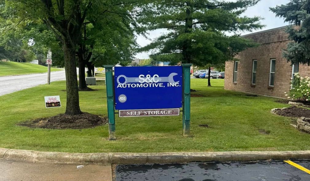 Sign for S&O Automotive, Inc., blue and white, with wrench graphic; in front of building, trees, and parking area  | S&C Automotive Inc