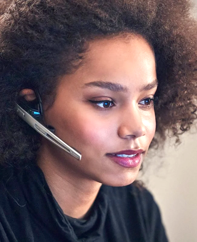 Woman with curly hair wearing a headset, looking slightly off-camera, possibly in a workspace.