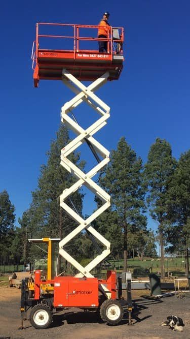 A person on an orange scissor lift. The lift is fully extended. A sunny day with a forest backdrop.