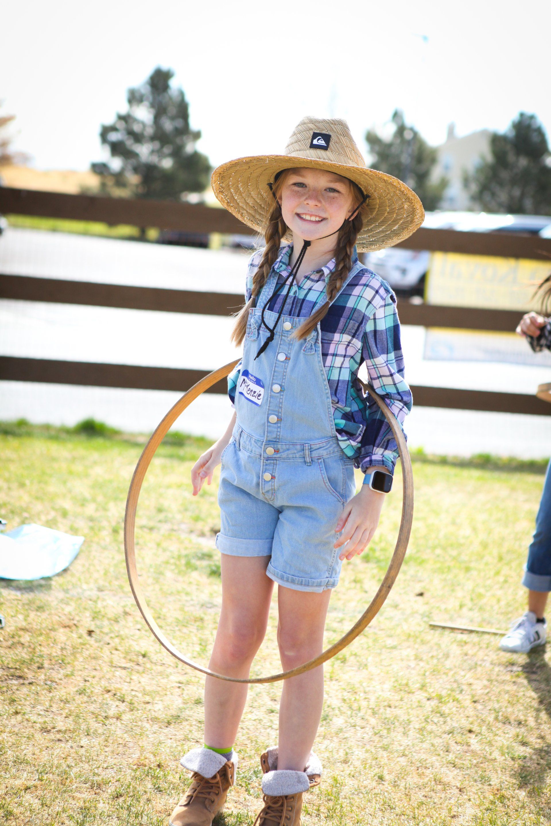 A young girl wearing a straw hat and overalls is holding a hula hoop.