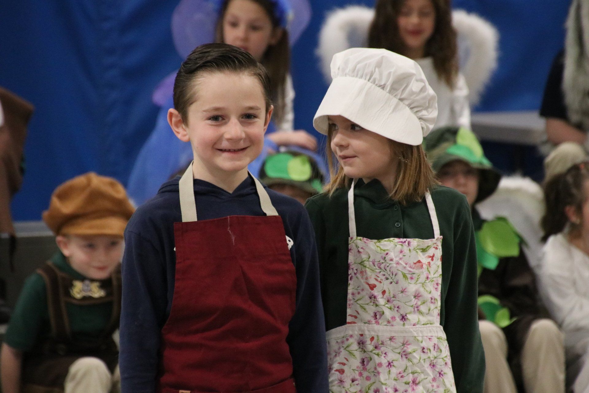 A boy and a girl wearing aprons and chef hats are standing next to each other.