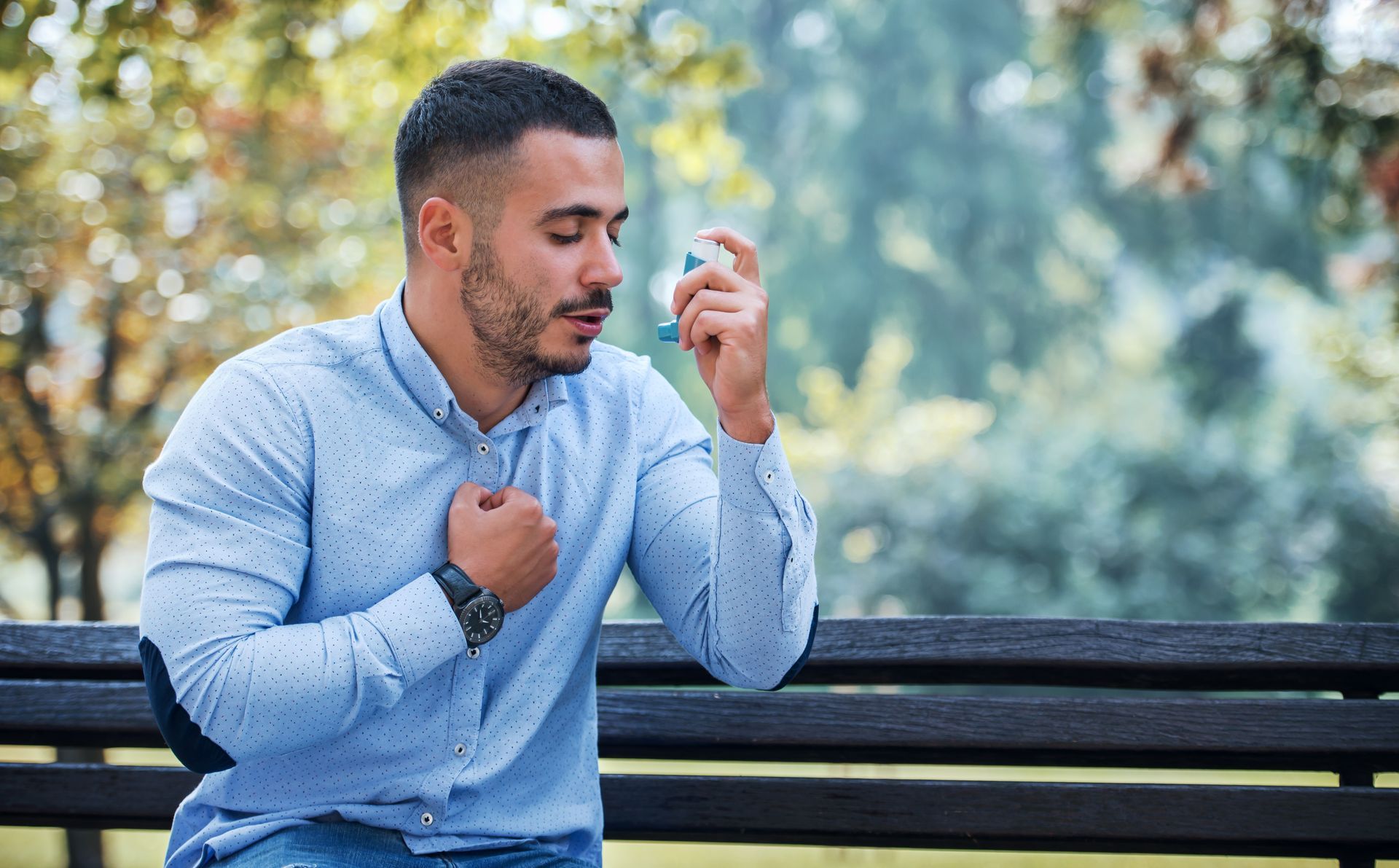 Young man treating asthma with inhaler sitting on a bench at a park