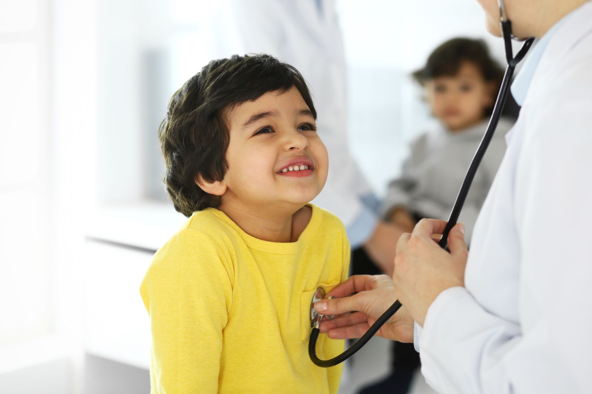 young boy wearing a yellow shirt smiling while the doctor holds a stethoscope to his chest