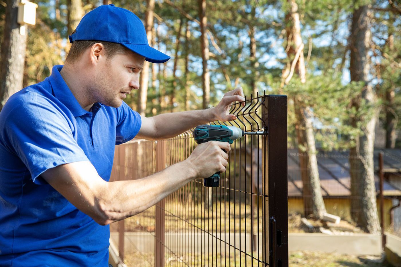 A fence contractor wearing a blue uniform, installing a welded metal mesh fence.