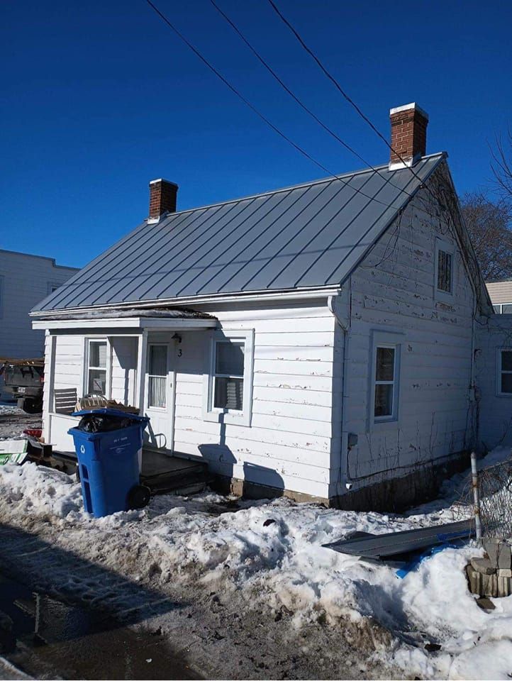 Une petite maison blanche avec un toit en métal est entourée de neige