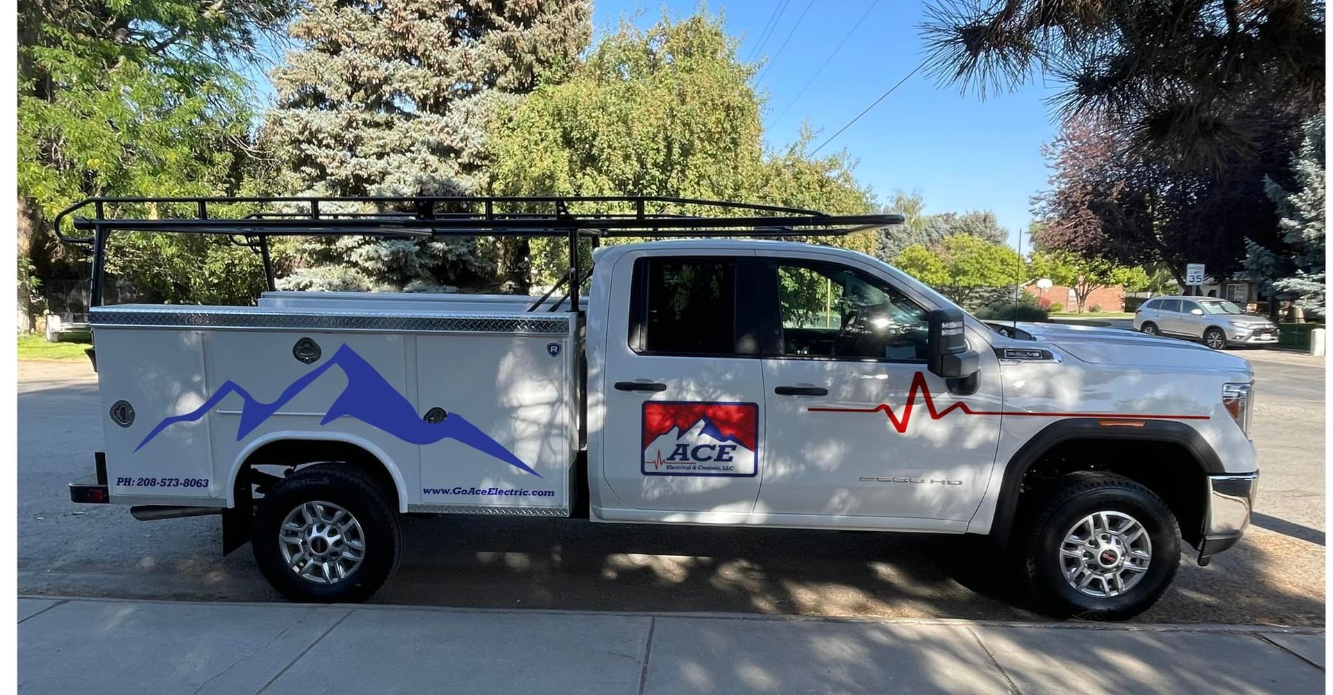 White work truck with mountain logo, toolboxes, and roof rack parked on a sidewalk.