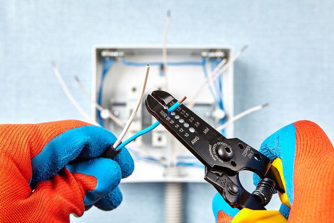 Hands wearing gloves strip wire with a tool, electrical box in the background.
