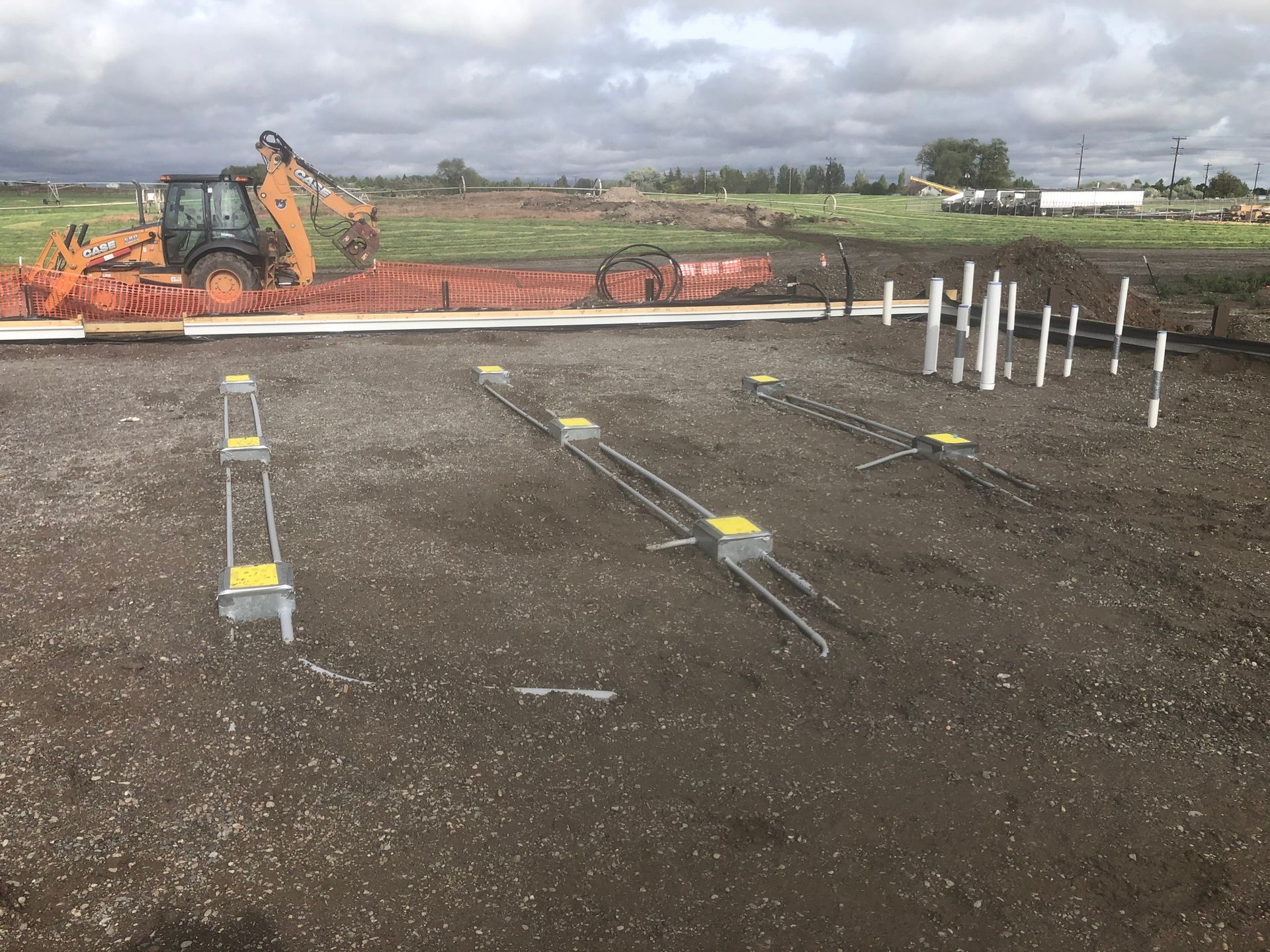 Construction site with a backhoe, exposed plumbing pipes, and metal supports on gravel. Overcast sky.