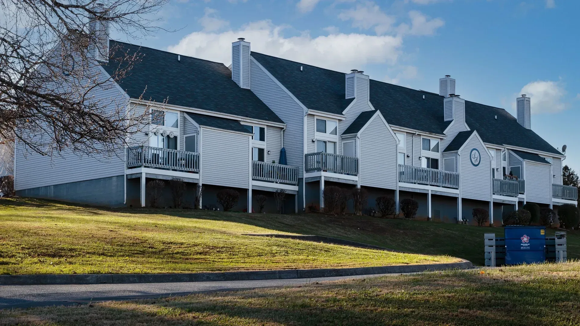 White multi-unit houses on a green hill with balconies and dark rooftops on a sunny day.