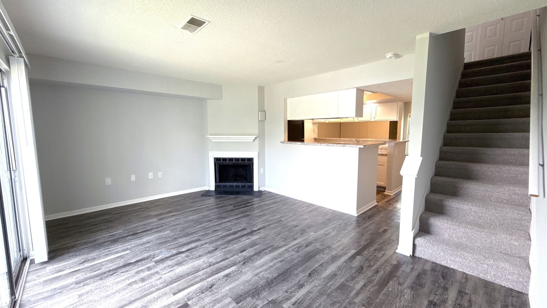 Empty living room with fireplace, stairs, and open kitchen. Gray wood-look floors and light walls.