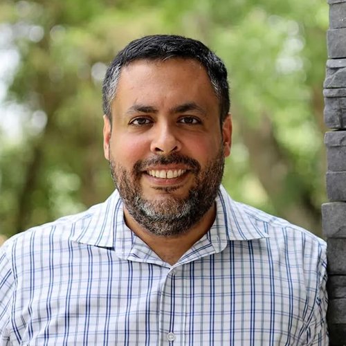 A smiling man with a beard and short dark hair wears a blue and white checkered shirt against an outdoor background.
