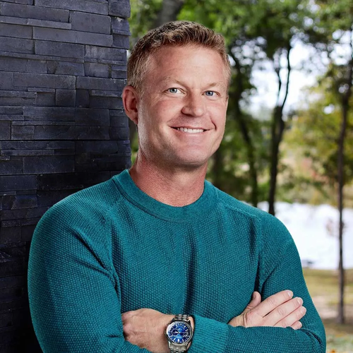 A person wearing a teal sweater smiles with their arms crossed, standing against a dark stone wall and natural backdrop.