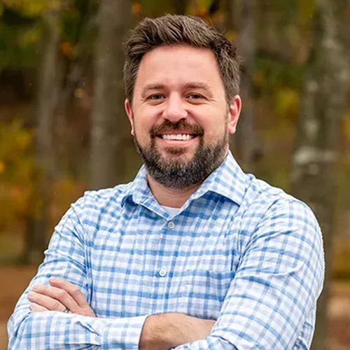 A person with a beard smiling with arms crossed, wearing a light blue and white gingham shirt in an outdoor setting.