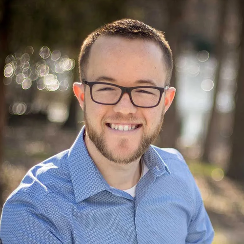 A smiling person with a beard and dark-rimmed glasses, wearing a blue button-down shirt outdoors with a blurred background.