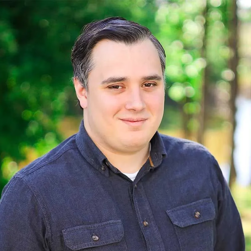 A person with short, dark hair wears a navy blue button-down shirt, smiling gently outdoors with a blurred green background.