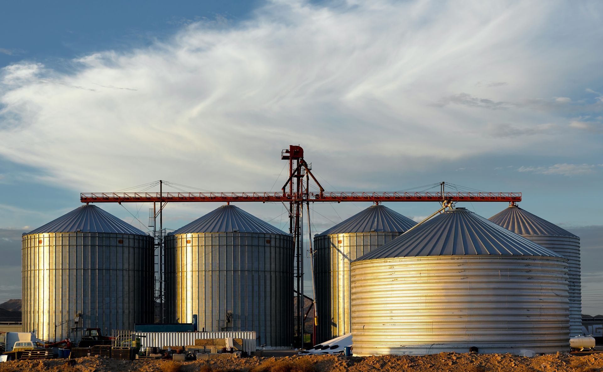 A row of shiny metal grain silos under a blue sky with streaky clouds, connected by a red overhead mechanical conveyor.