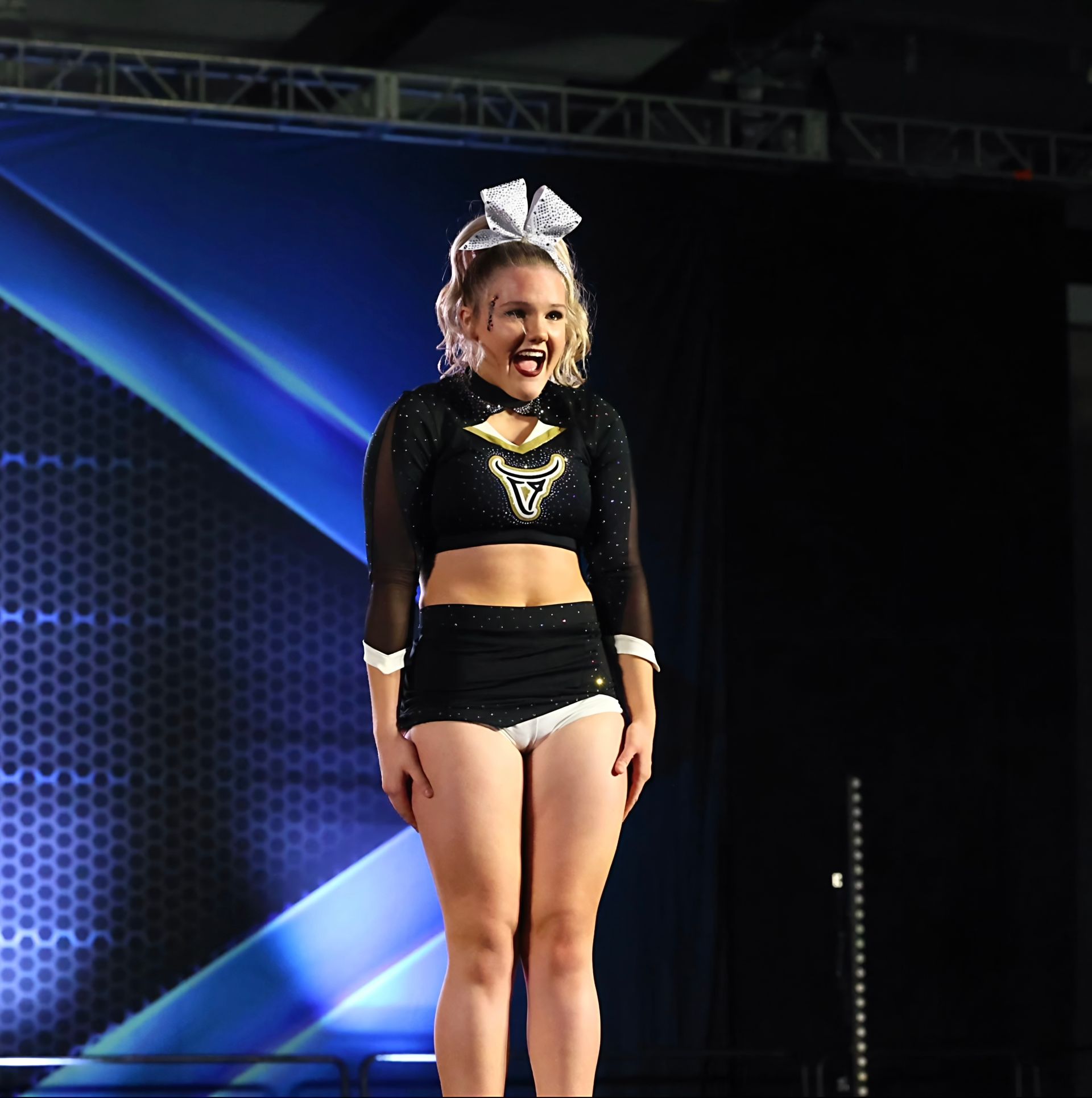 Cheerleader in black uniform on stage with a white bow, smiling, black background with blue streaks.
