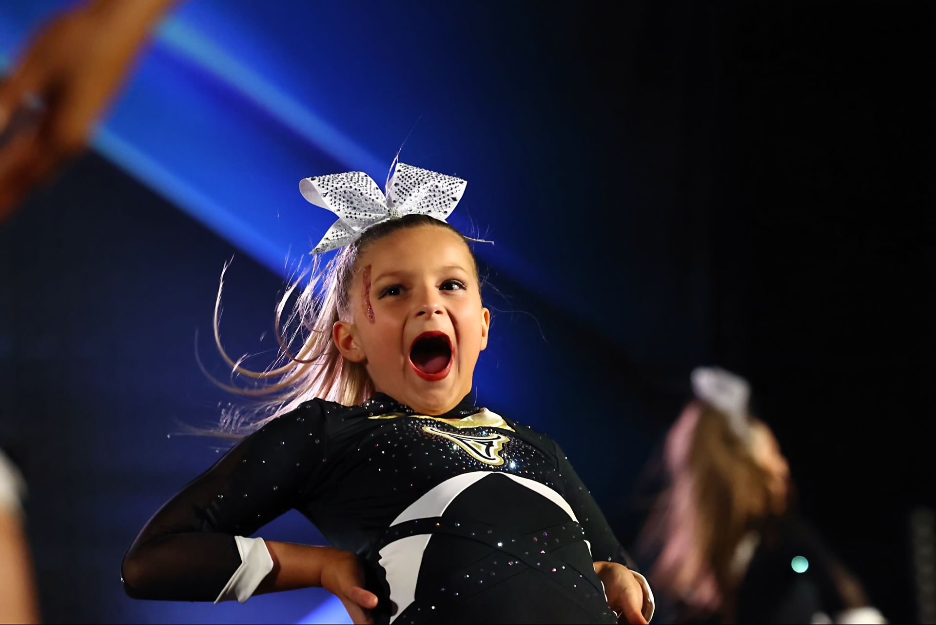 Young cheerleader with mouth open wide, wearing bow and black uniform with white accents.
