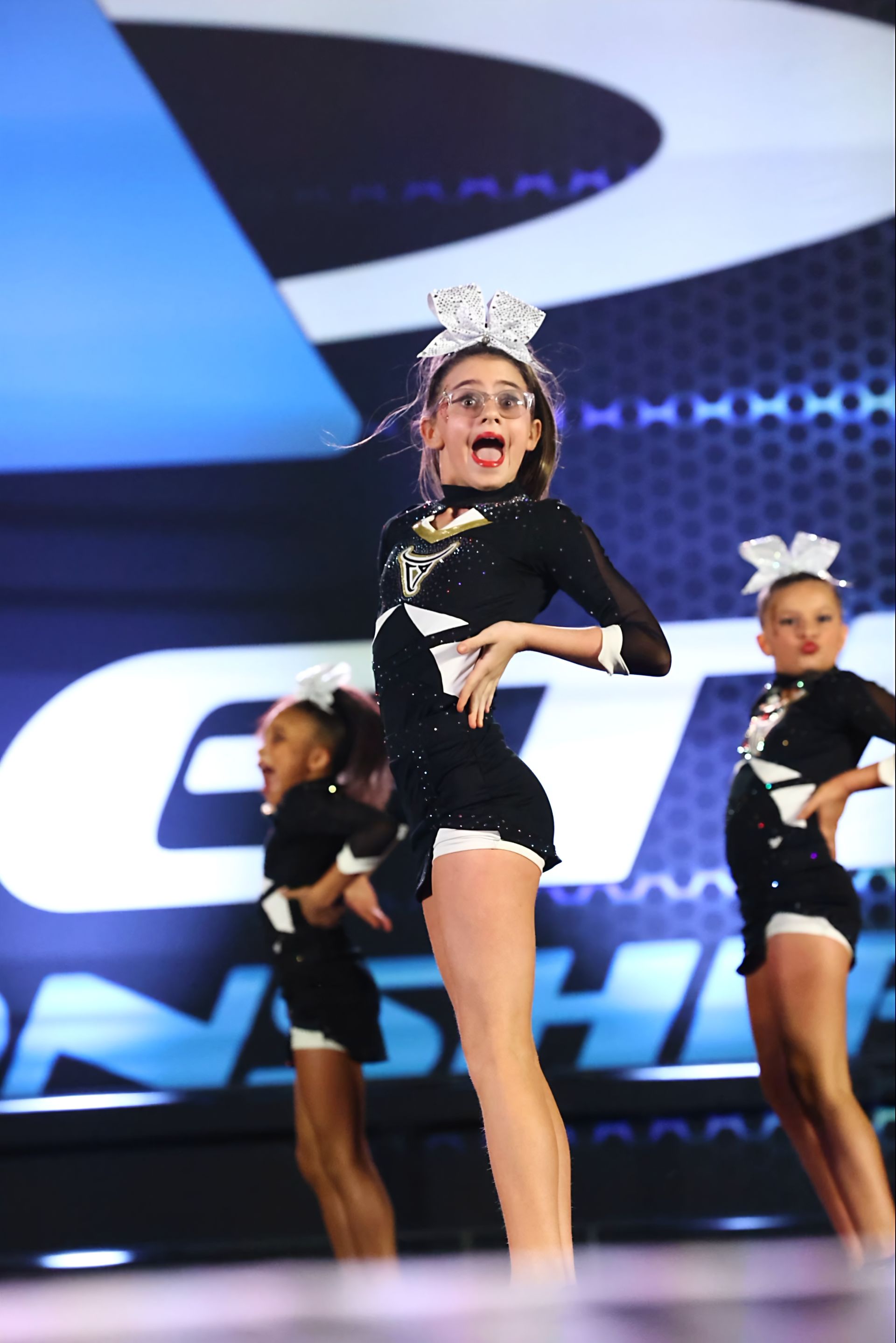 Cheerleaders in black and white uniforms perform on a stage. One girl is center, mid-leap, mouth open.