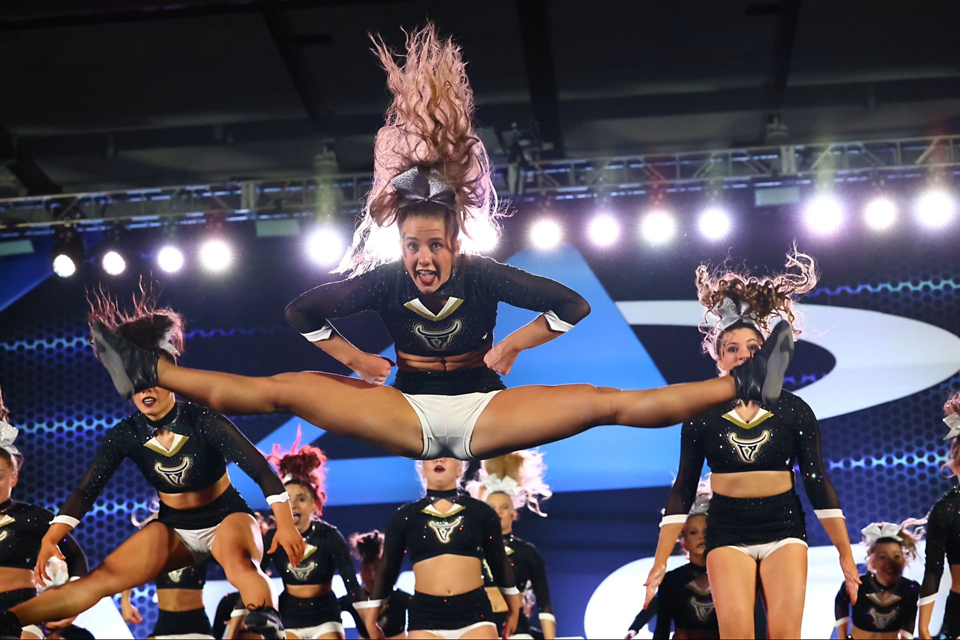 Cheerleader mid-air split jump; black and gold uniform; stage with lights; other cheerleaders present.