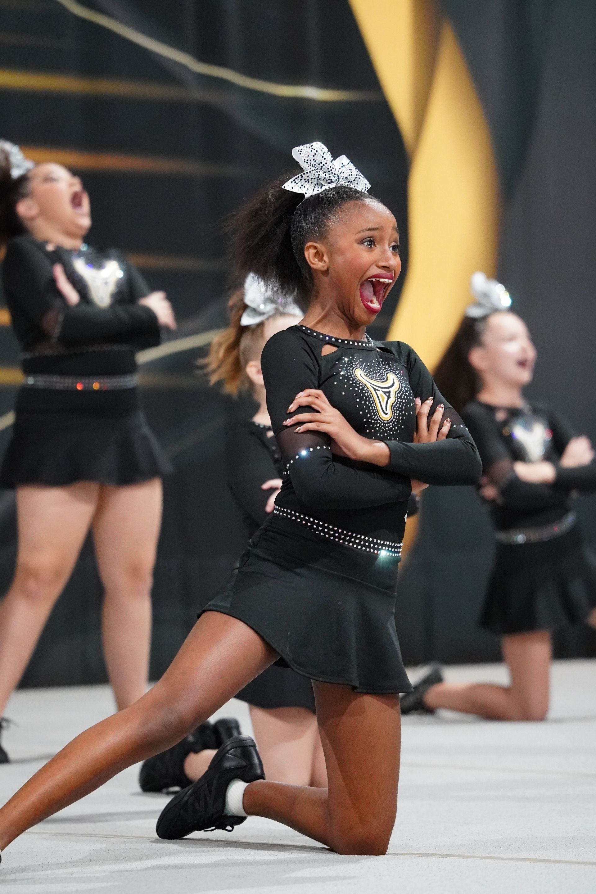 Cheerleaders in black outfits perform. A girl kneels, arms crossed, mouth open, looking excited.