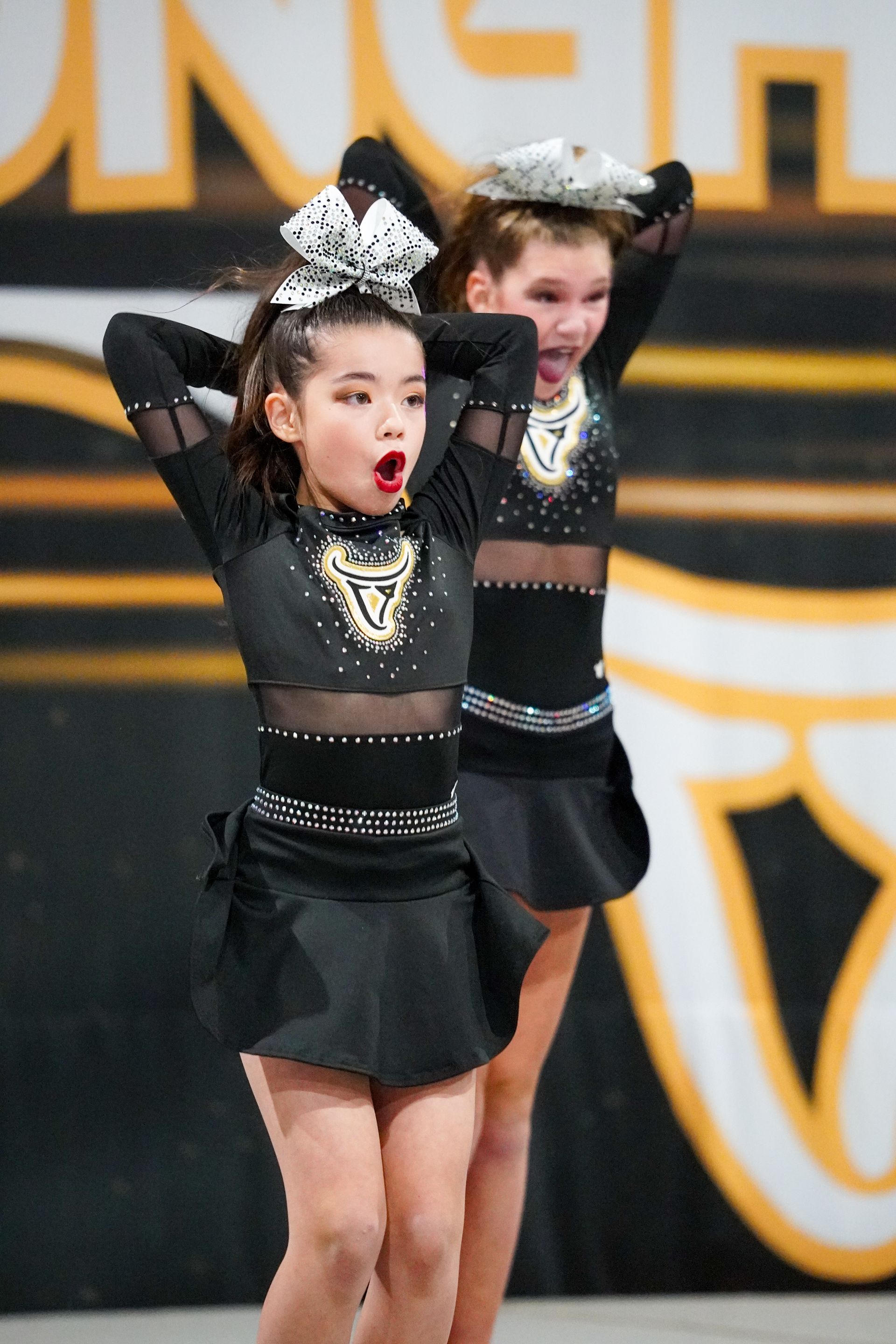 Two cheerleaders in black uniforms with arms overhead, performing on a stage.