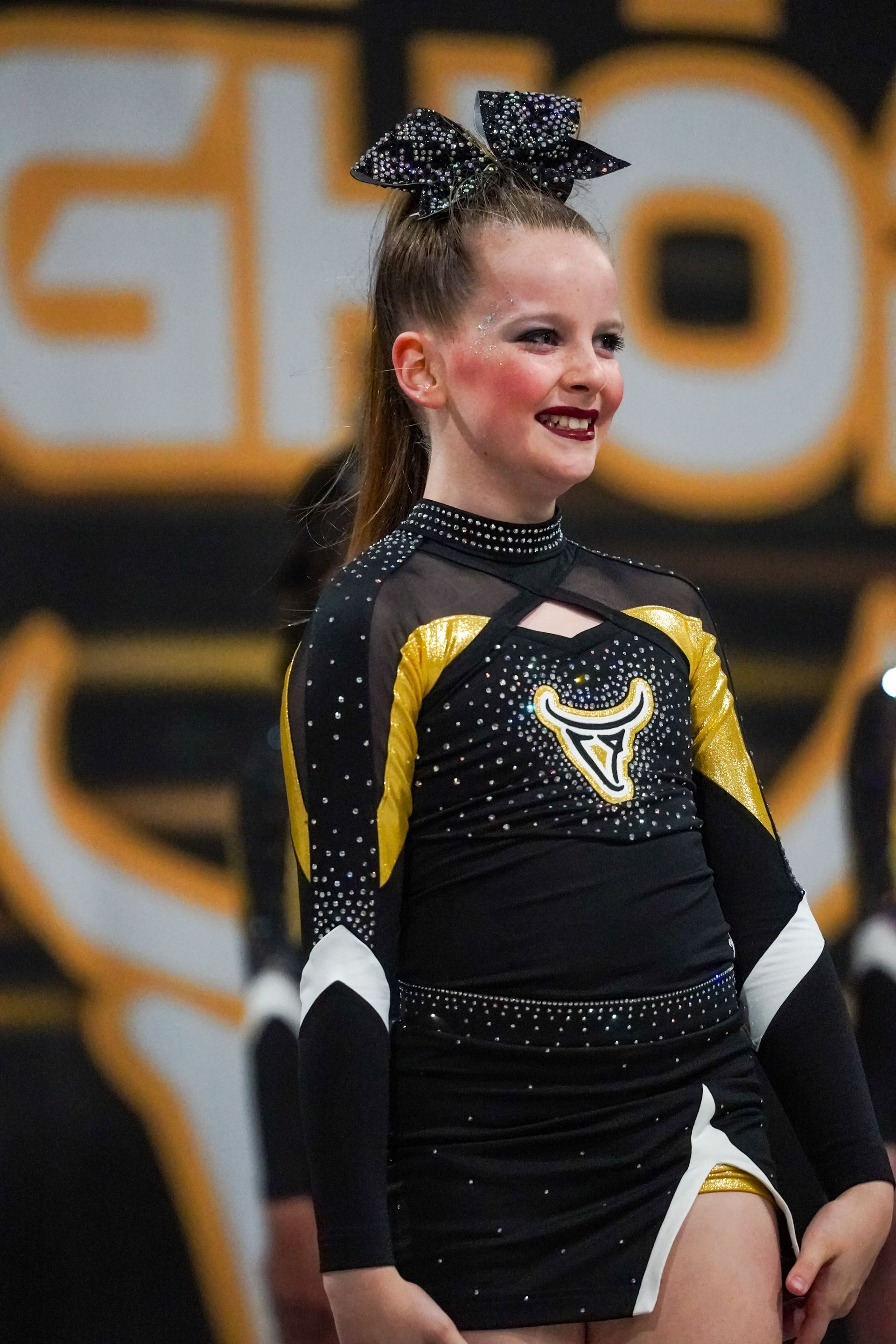 Young cheerleader in black and gold uniform smiles at competition.