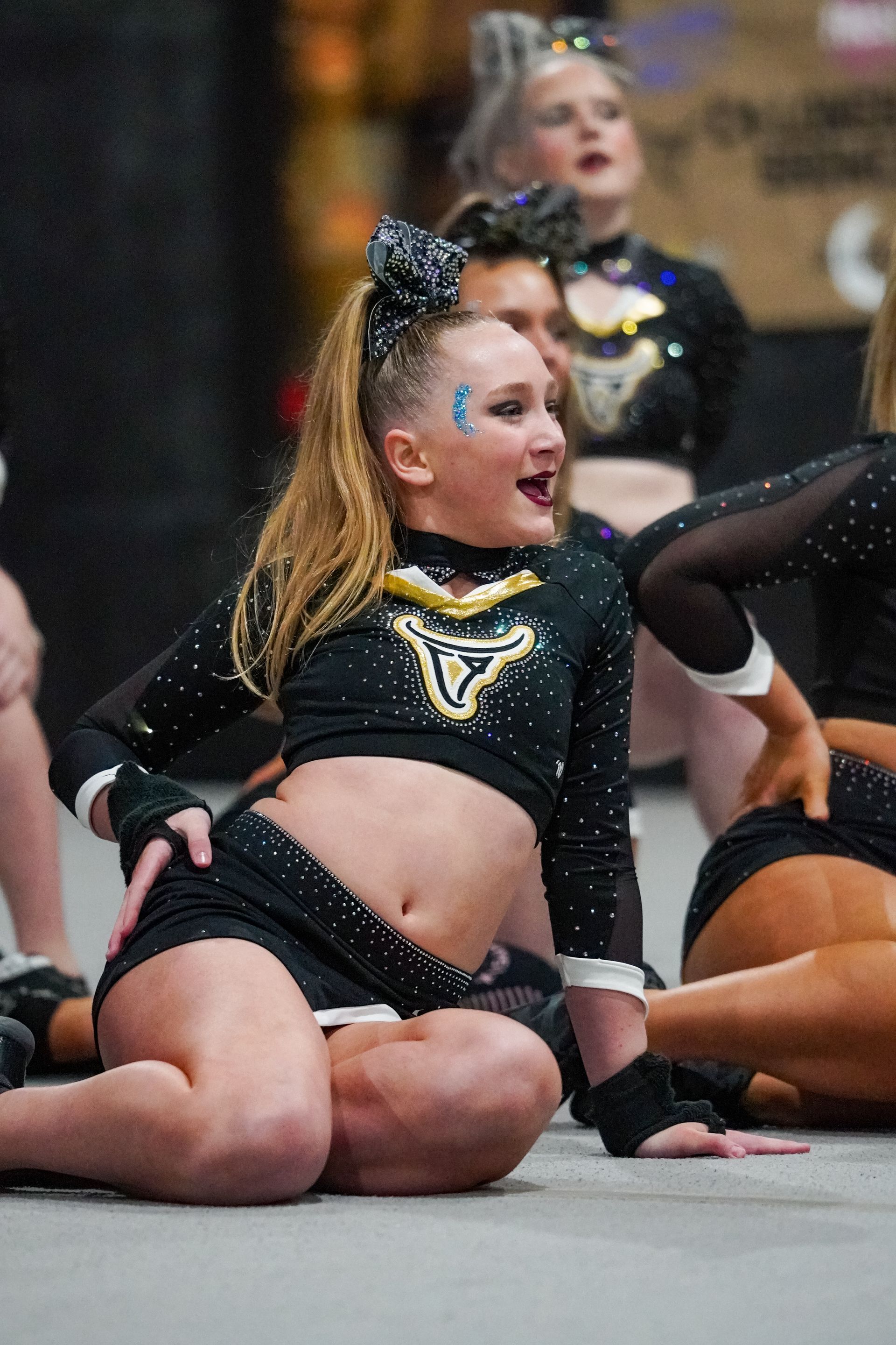 Cheerleader in a black uniform performing. She is smiling, with one hand on hip, in a gym.