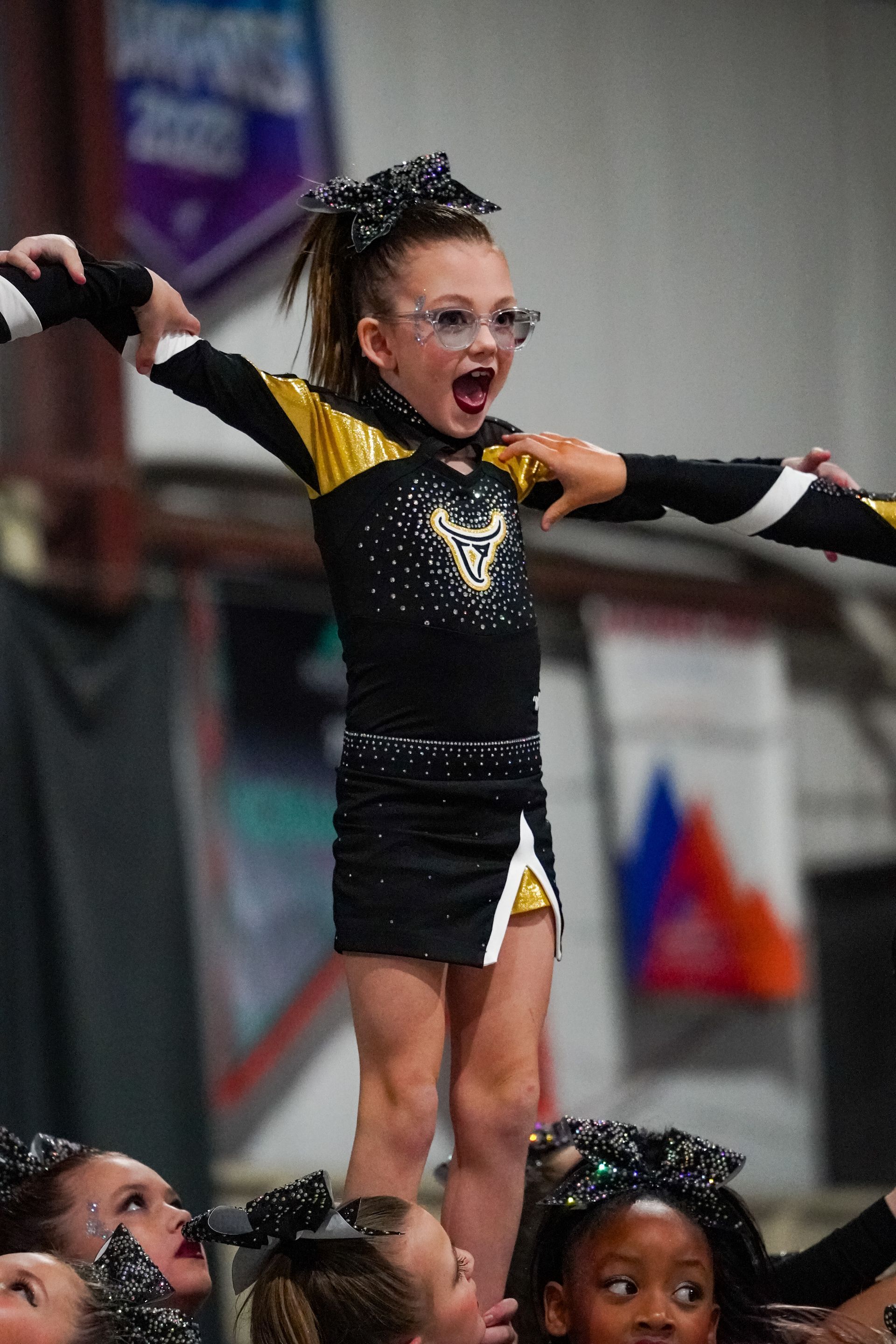 Young cheerleader in a black and gold uniform with arms raised, balanced atop teammates during a performance.
