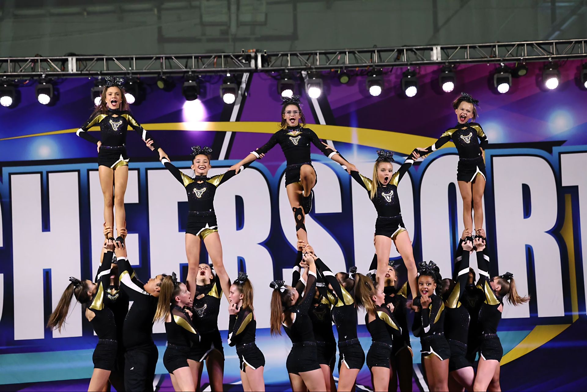 Cheerleaders performing a stunt at a competition. Black and gold uniforms, blue and purple background. "Cheersport" sign.