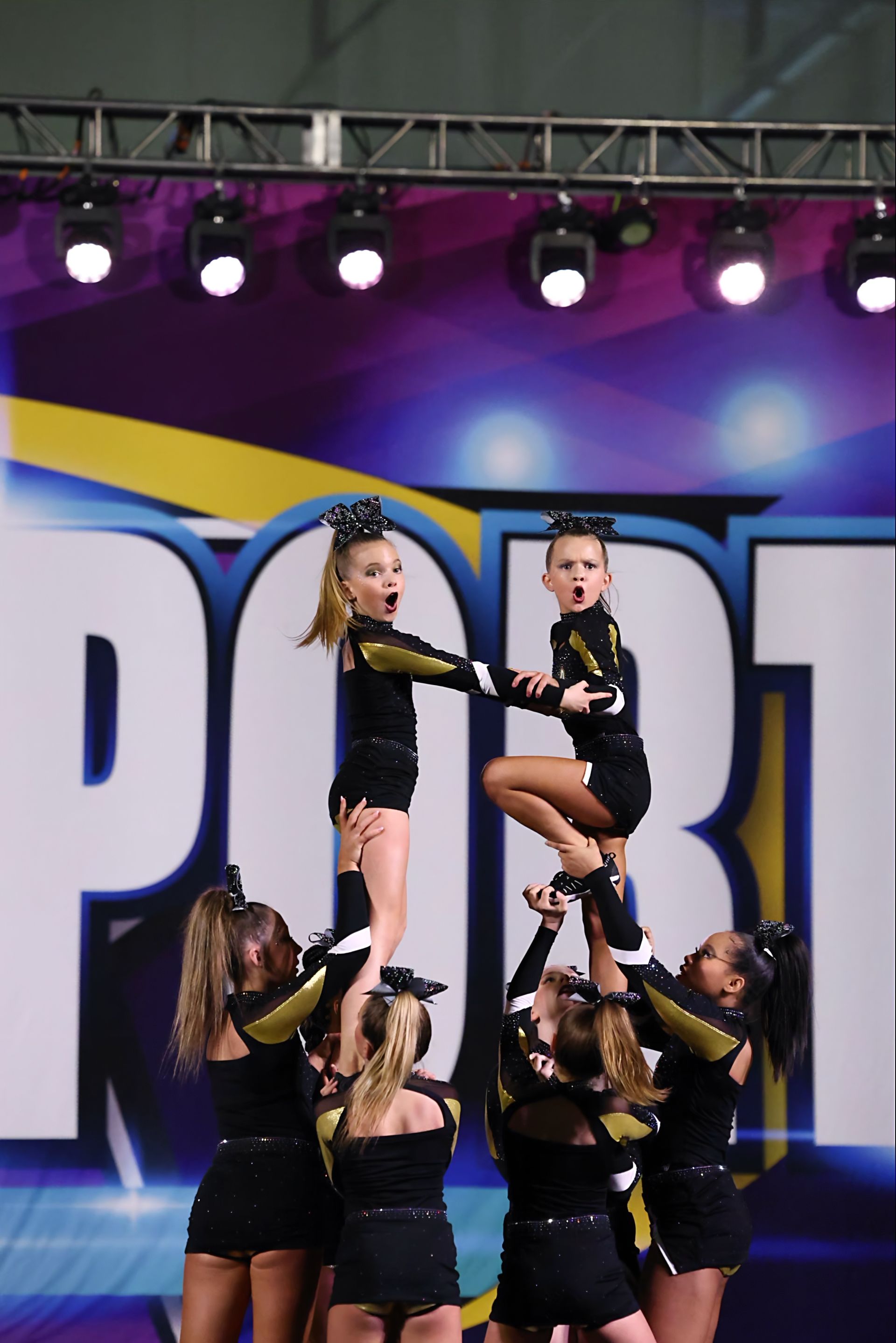 Cheerleaders performing a pyramid stunt indoors; black and gold uniforms; stage backdrop with the word 