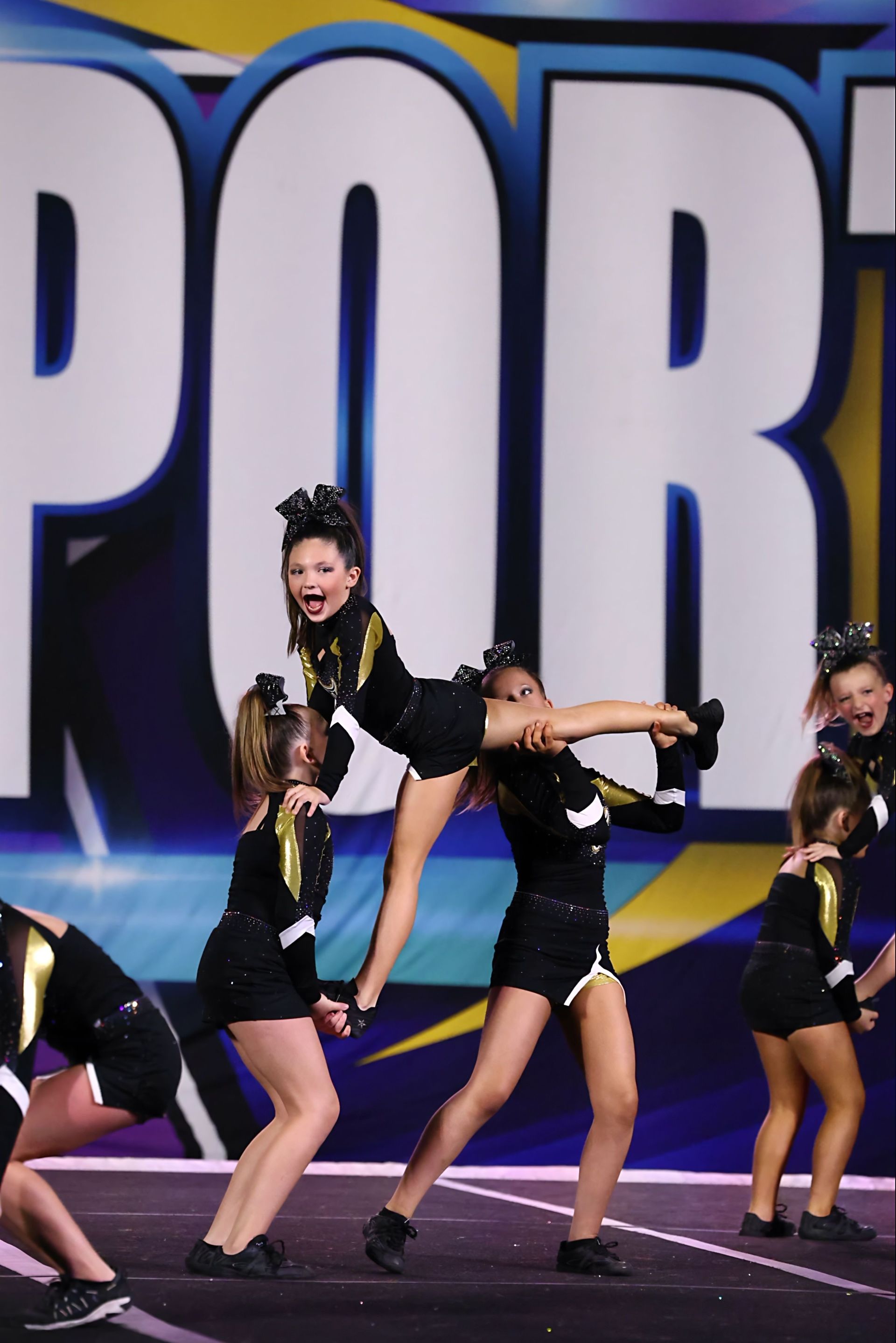 Cheerleaders in black and gold uniforms performing a stunt on a mat with 