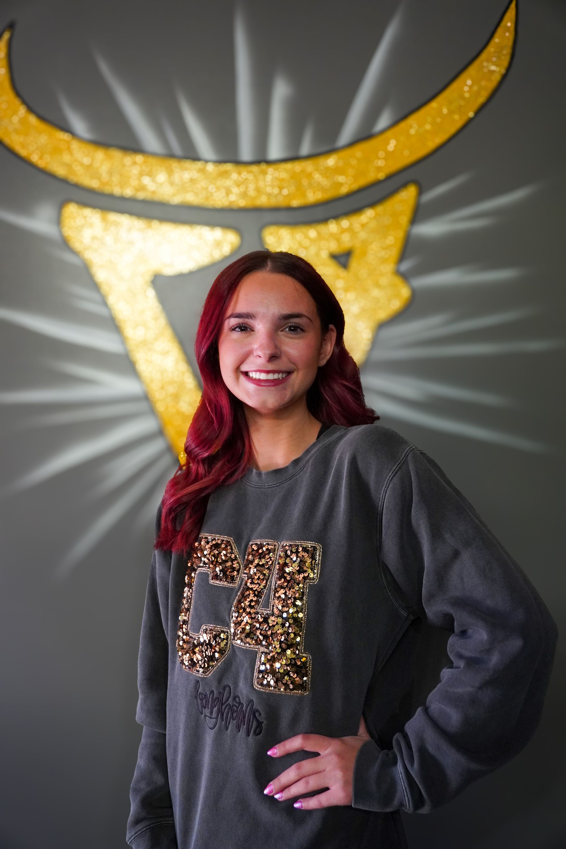Woman with red hair smiling, wearing a gray sweatshirt in front of a gold bull logo.