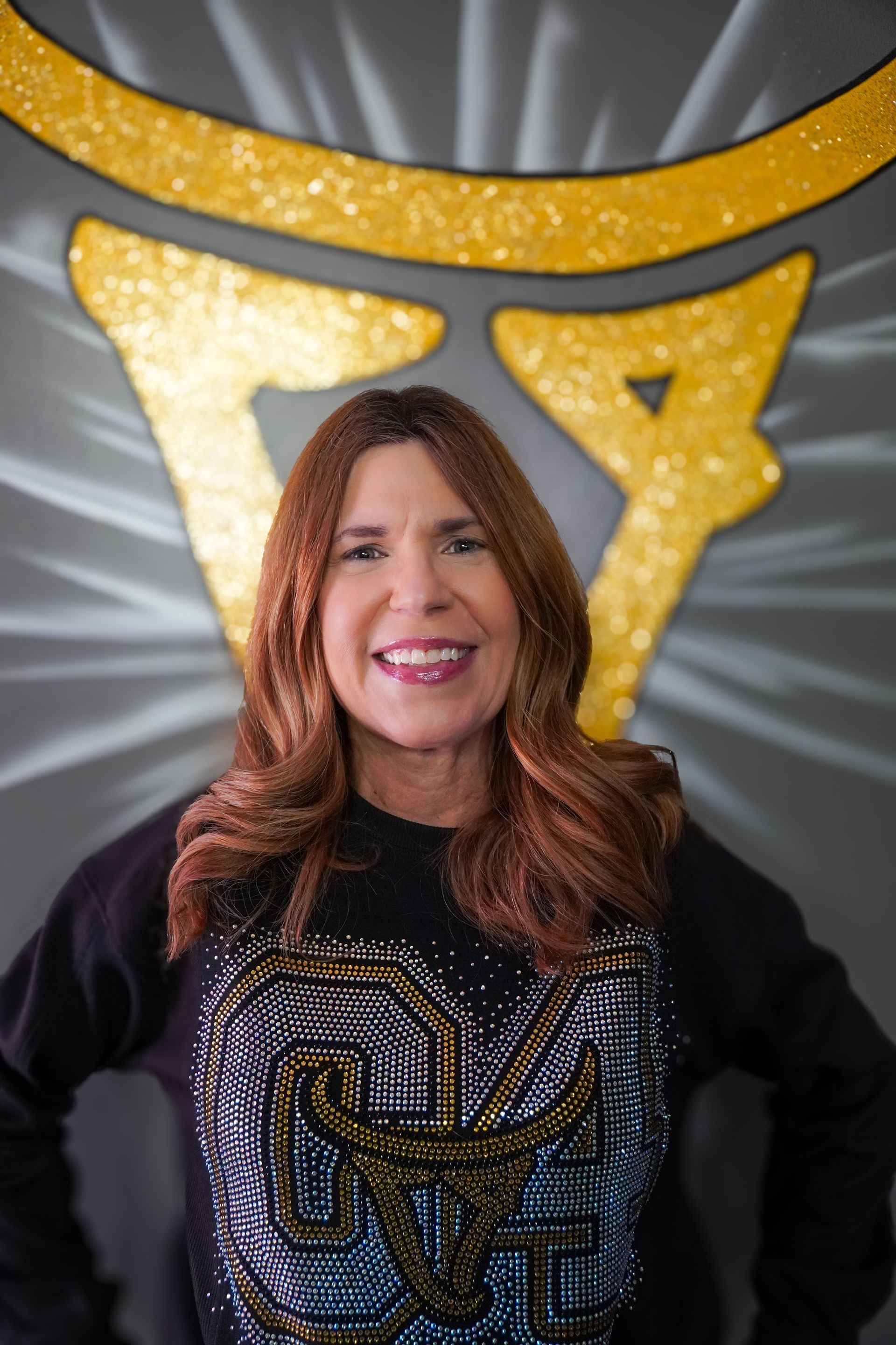 Woman smiling, posing in front of gold-painted symbol on a gray wall. She wears a black top with embellished design.
