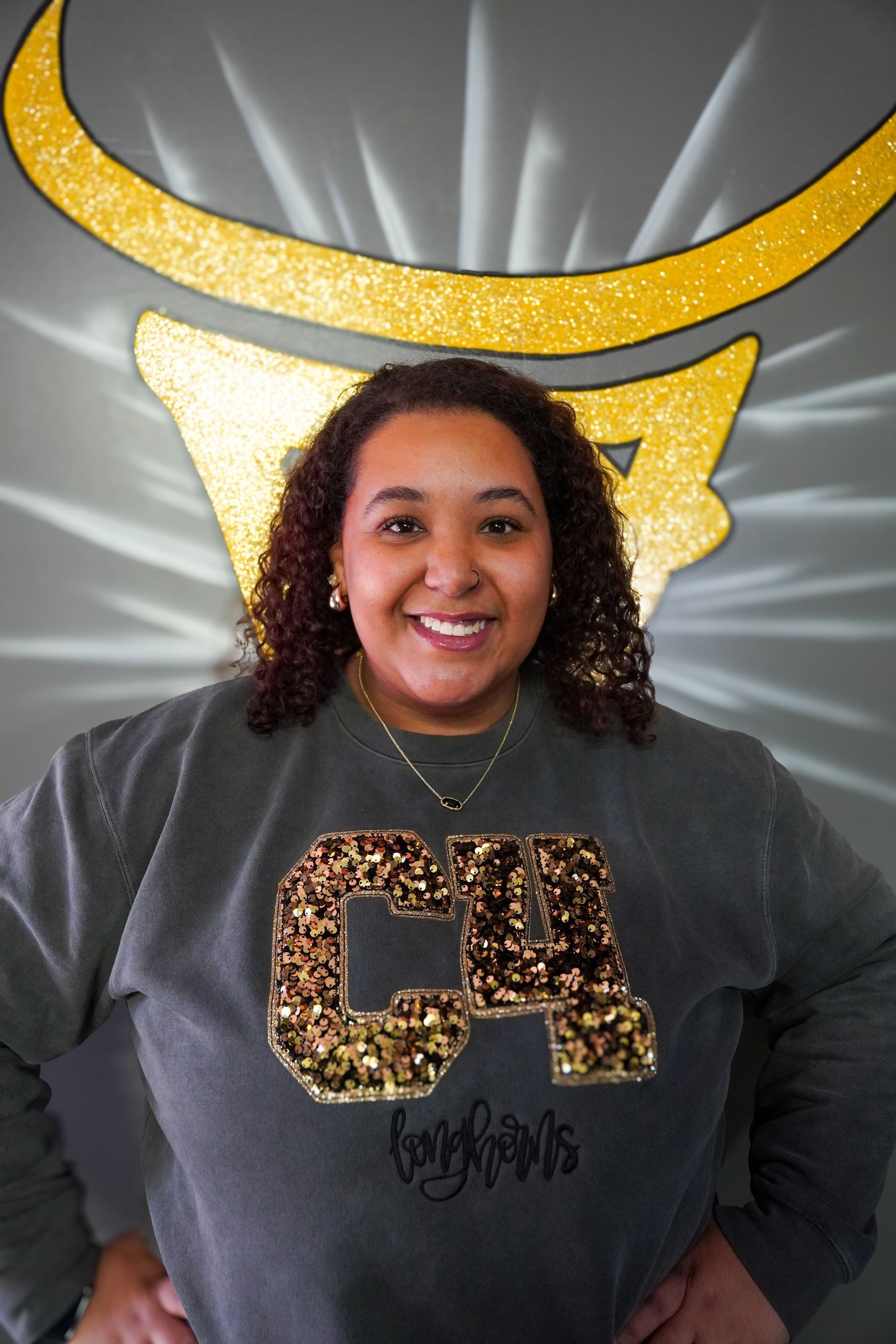 Woman smiling, wearing a gray sweatshirt with gold lettering, standing in front of a gold bull logo on a gray wall.